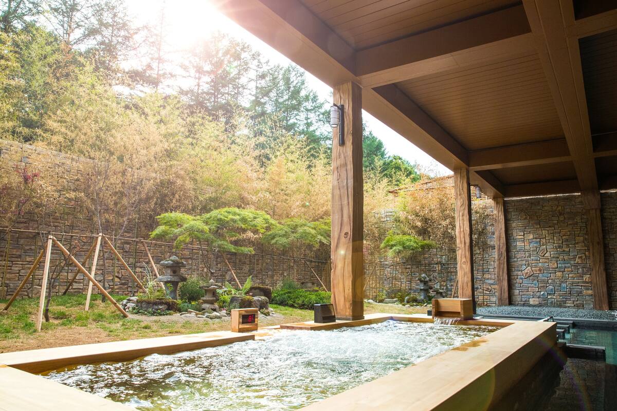 En traditionel japansk onsen med træterrasse, stenvæg og naturligt landskab.