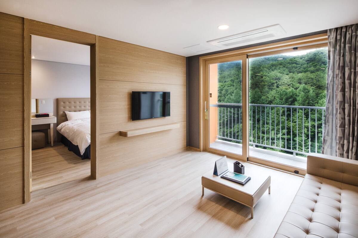 Et moderne hotelværelse med trægulv, et stort vindue med balkon, et fladskærms-tv monteret på væggen og en behagelig sofa.