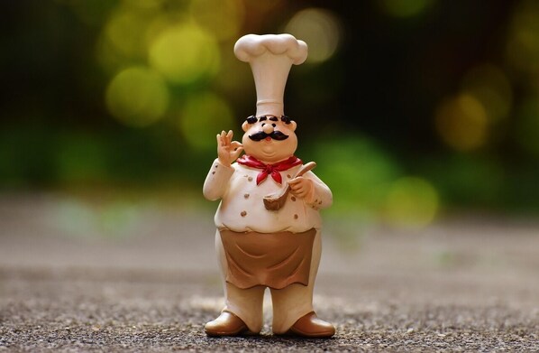 Una figurita de un cocinero con un sombrero blanco alto, un lazo rojo y un delantal marrón, de pie sobre una superficie con textura.