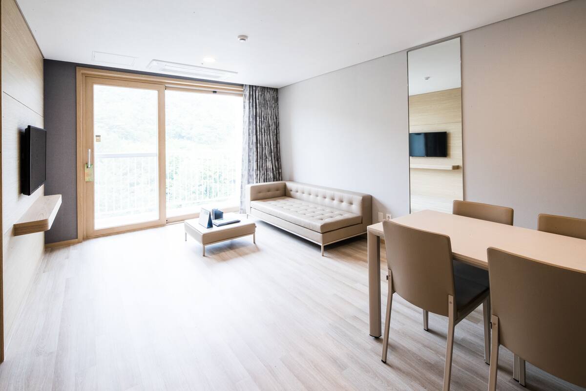 En moderne stue med sofa, spisebord med stole, fjernsyn ophængt på væggen og en skydedør, der fører ud til en balkon.