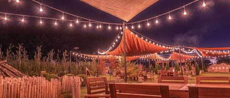 Un área de estar al aire libre con bancos y mesas de madera, luces colgantes y un toldo.