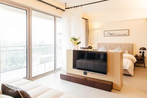 A modern bedroom with a large TV, a bed, a bedside table, and a window with a view of the outdoors.