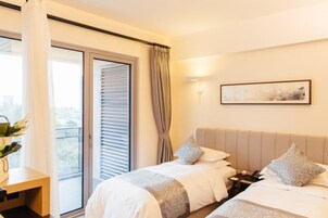 A hotel room with two beds, a large window with blinds, a desk with a plant, and a painting on the wall.