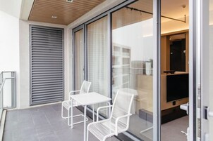 A modern balcony with a glass door, white metal chairs, and a flat-screen TV.
