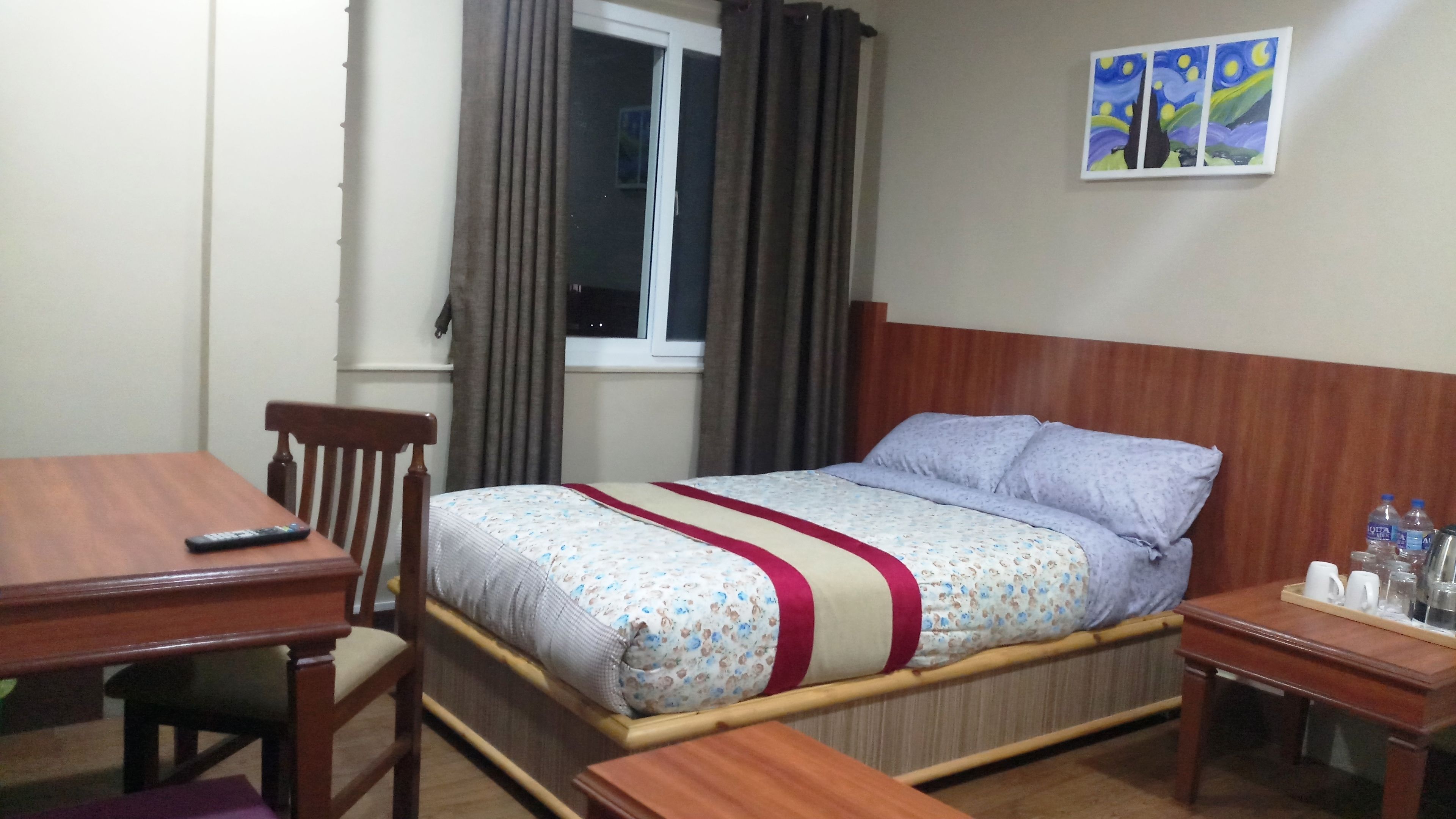 Een hotelkamer met een houten bed, een houten bureau, een stoel, een nachtkastje met waterflessen en een raam met gordijnen.