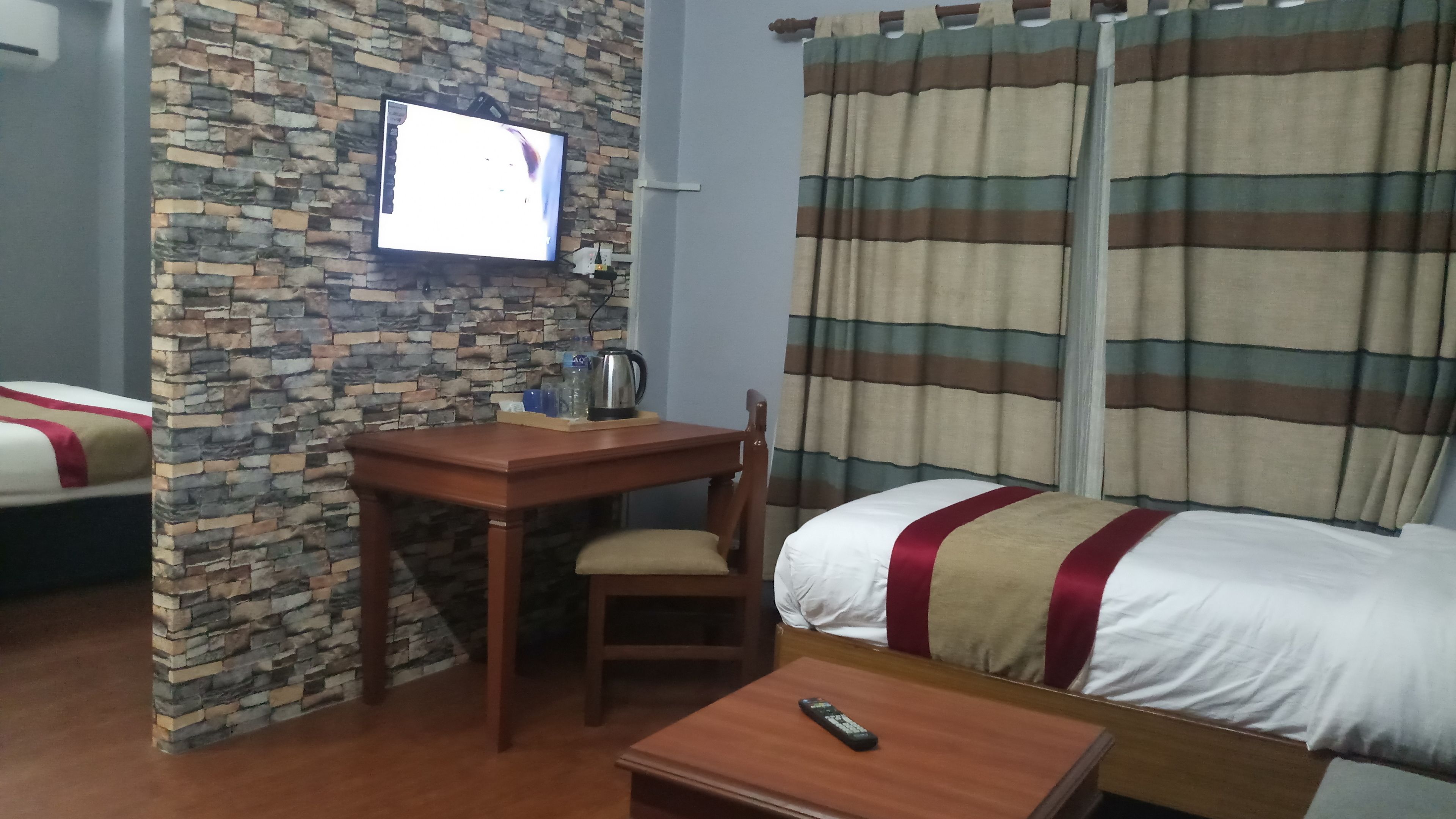 A hotel room with a stone wall, a wooden desk with a chair, a bed with striped curtains, and a television mounted on the wall.