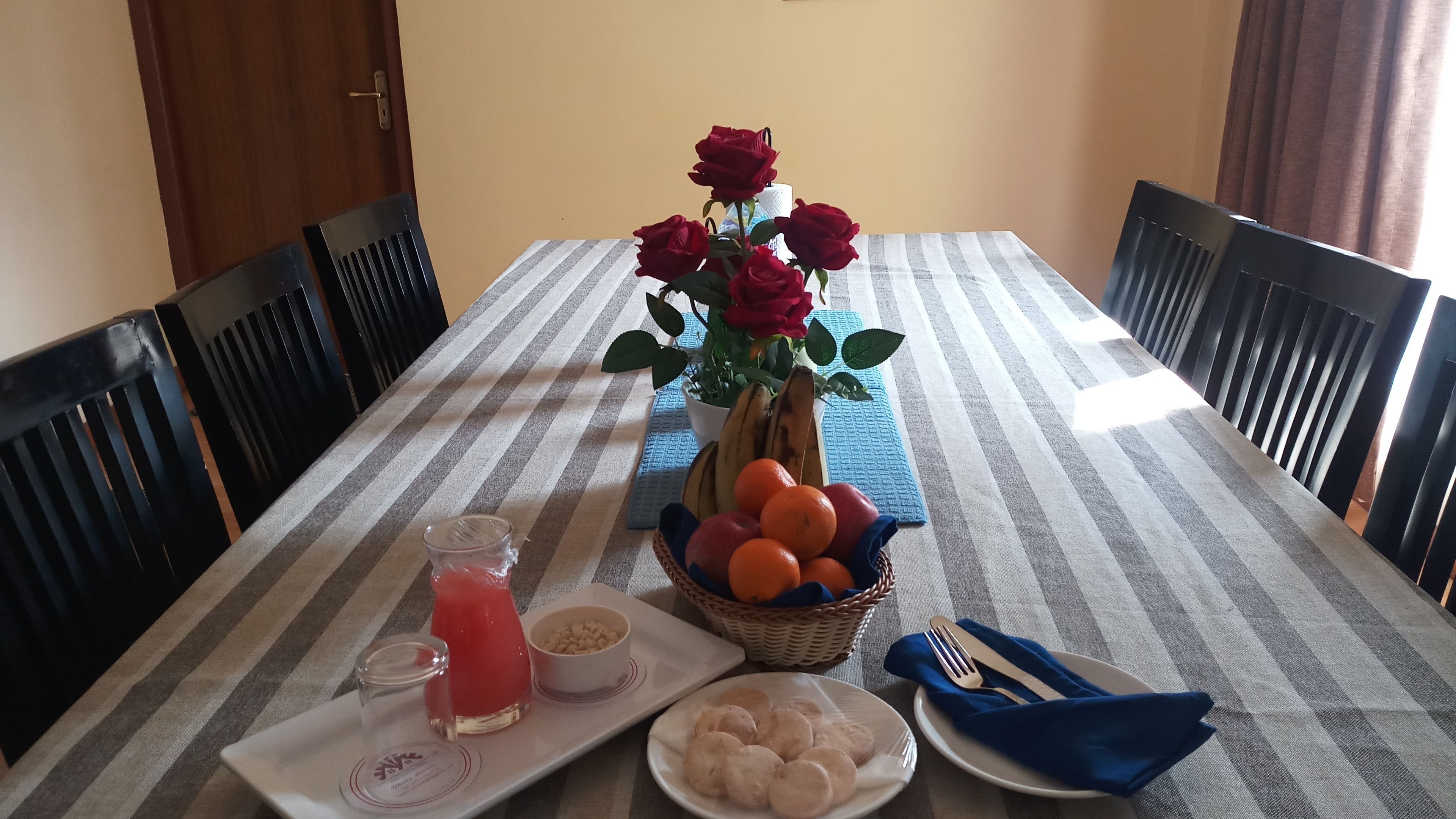 A dining table with a striped tablecloth, a vase of red roses, a basket of oranges, a plate of biscuits, a glass of pink drink, and a bowl of nuts.