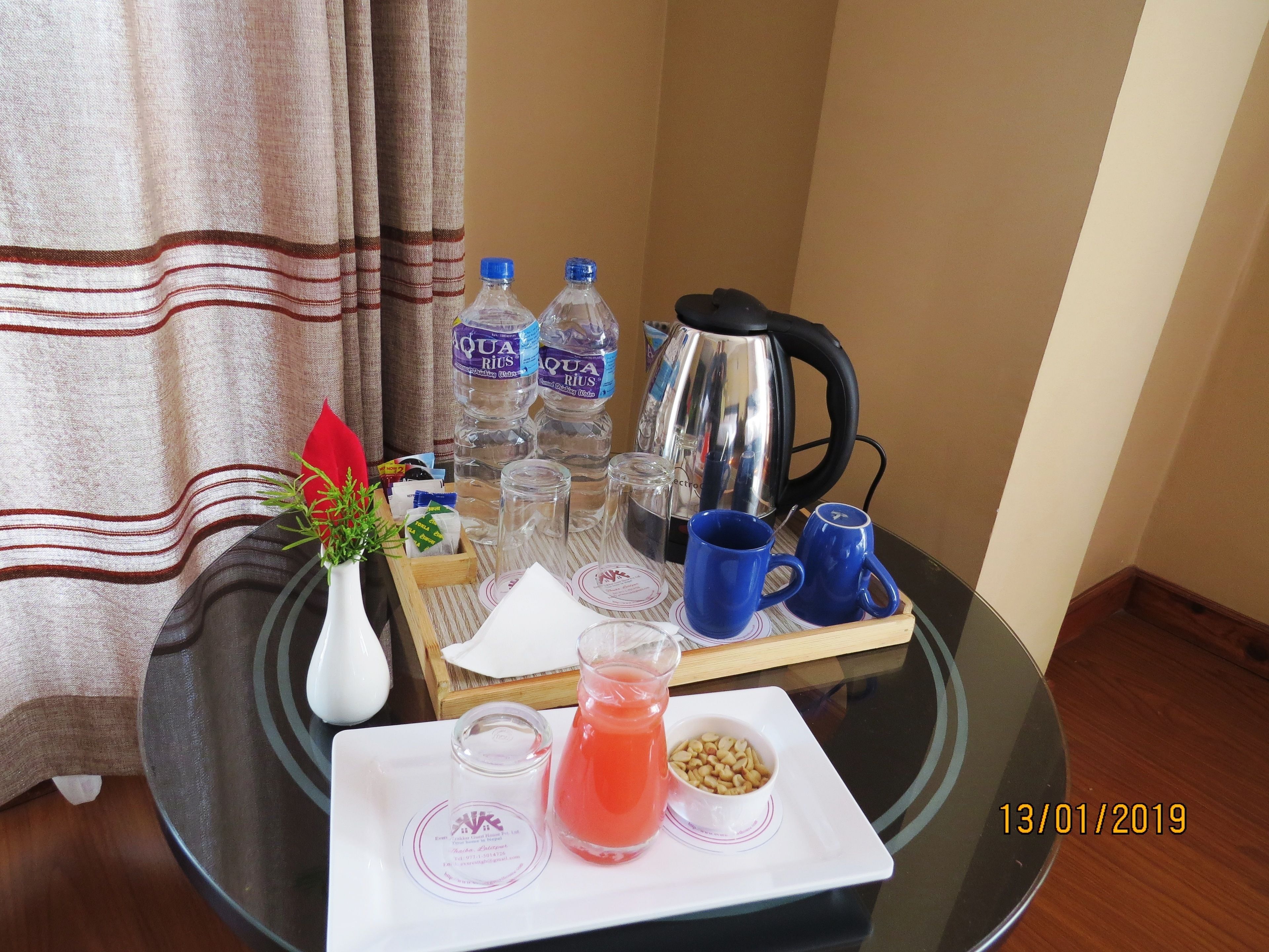 A breakfast tray with water bottles, a kettle, blue mugs, a pitcher of orange juice, a small bowl of nuts, and a white napkin on a glass table.