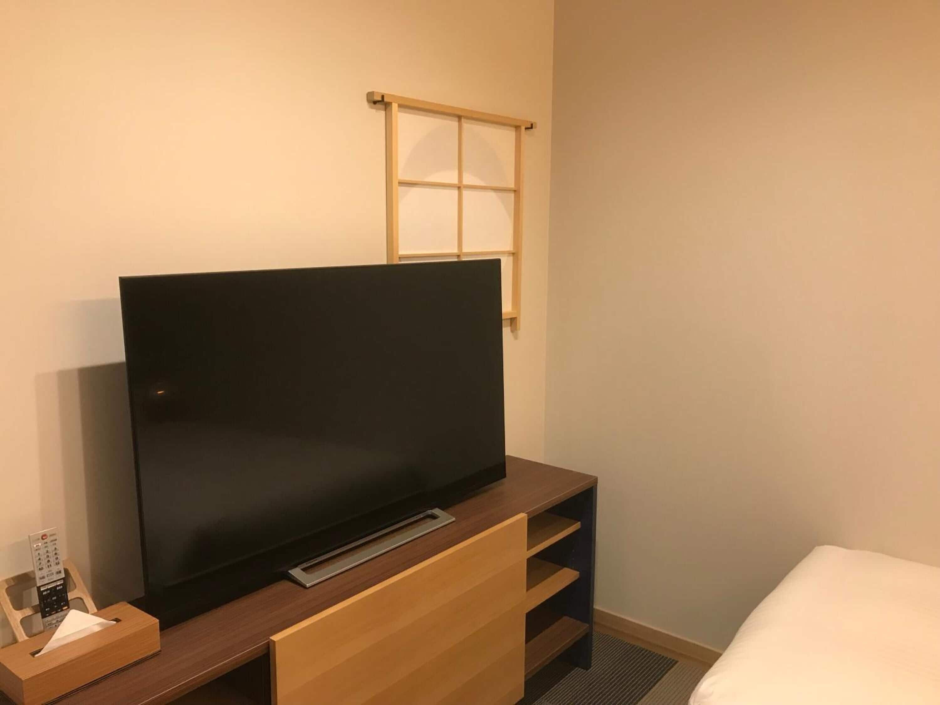 A hotel room with a flat-screen TV on a wooden stand, a bedside table with a remote control, and a window with a wooden blind.