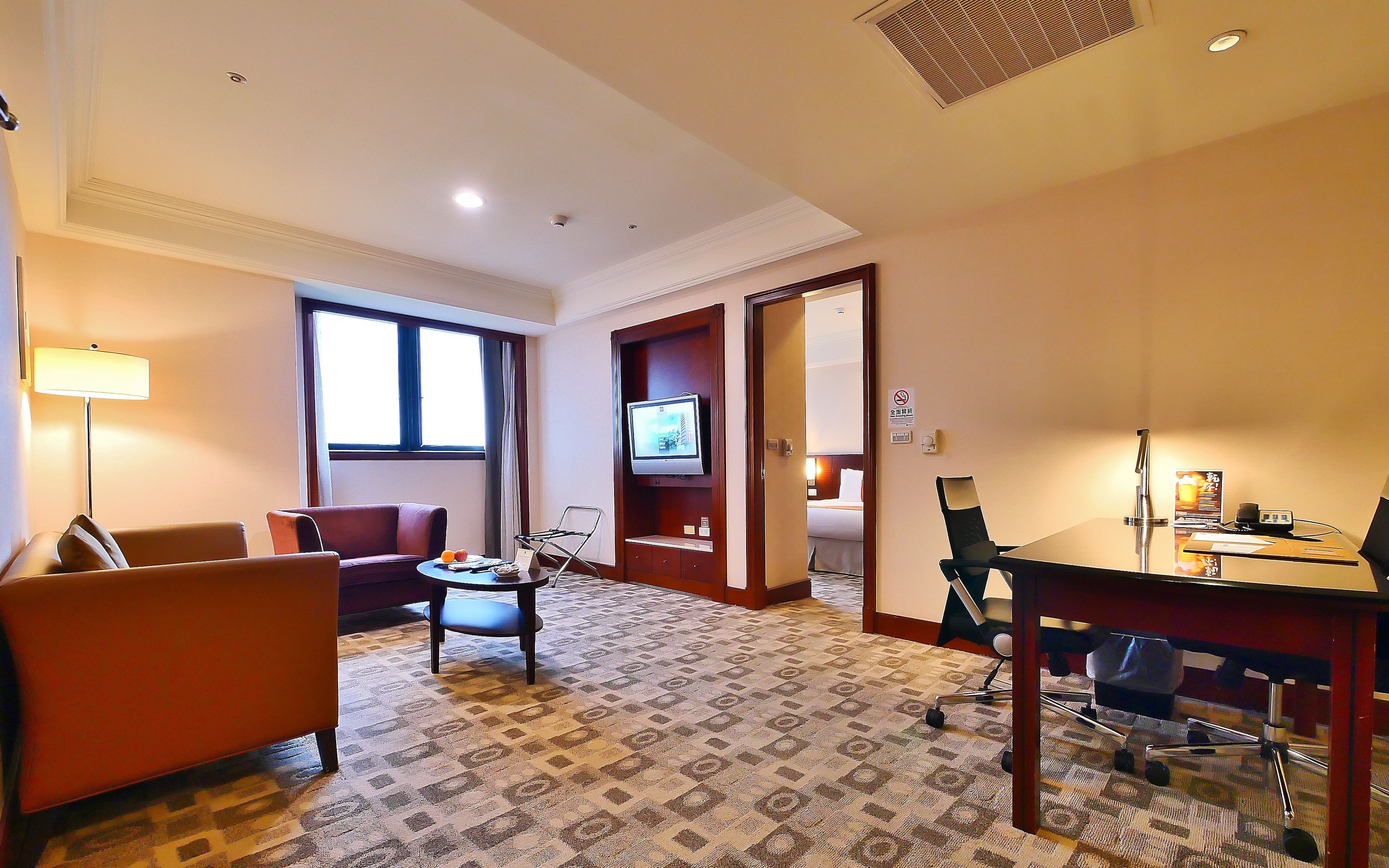 Una habitación de hotel con sofá, sillón, televisor, escritorio y una alfombra estampada.