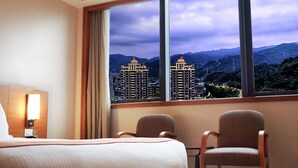 Una habitación de hotel con un ventanal grande, dos sillas, una cama y una mesita de noche con lámpara.
