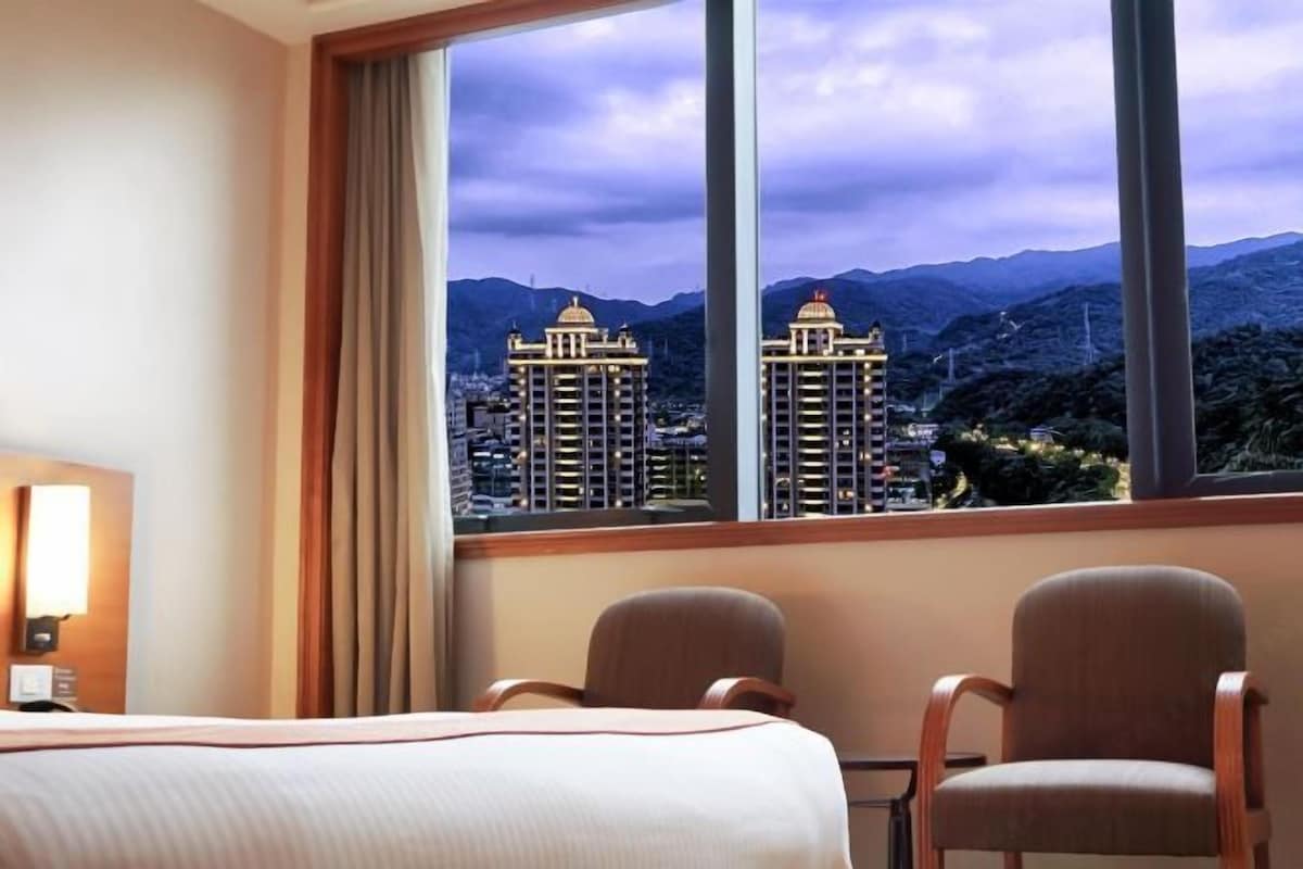 A hotel room with a large window offering a view of a cityscape and mountains.