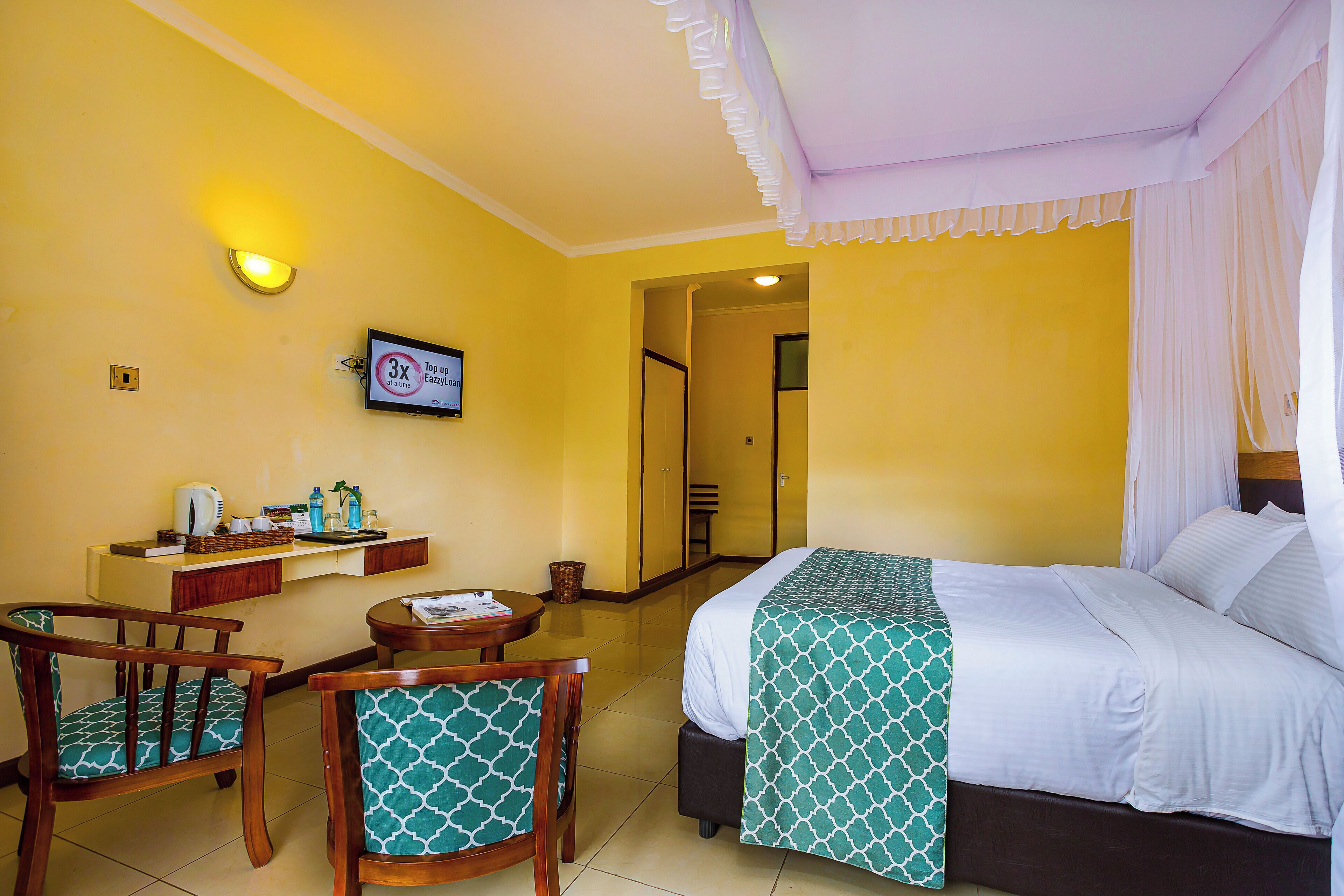 Habitación de hotel con cama, dos sillas, una mesita, estante con artículos de aseo y televisor.