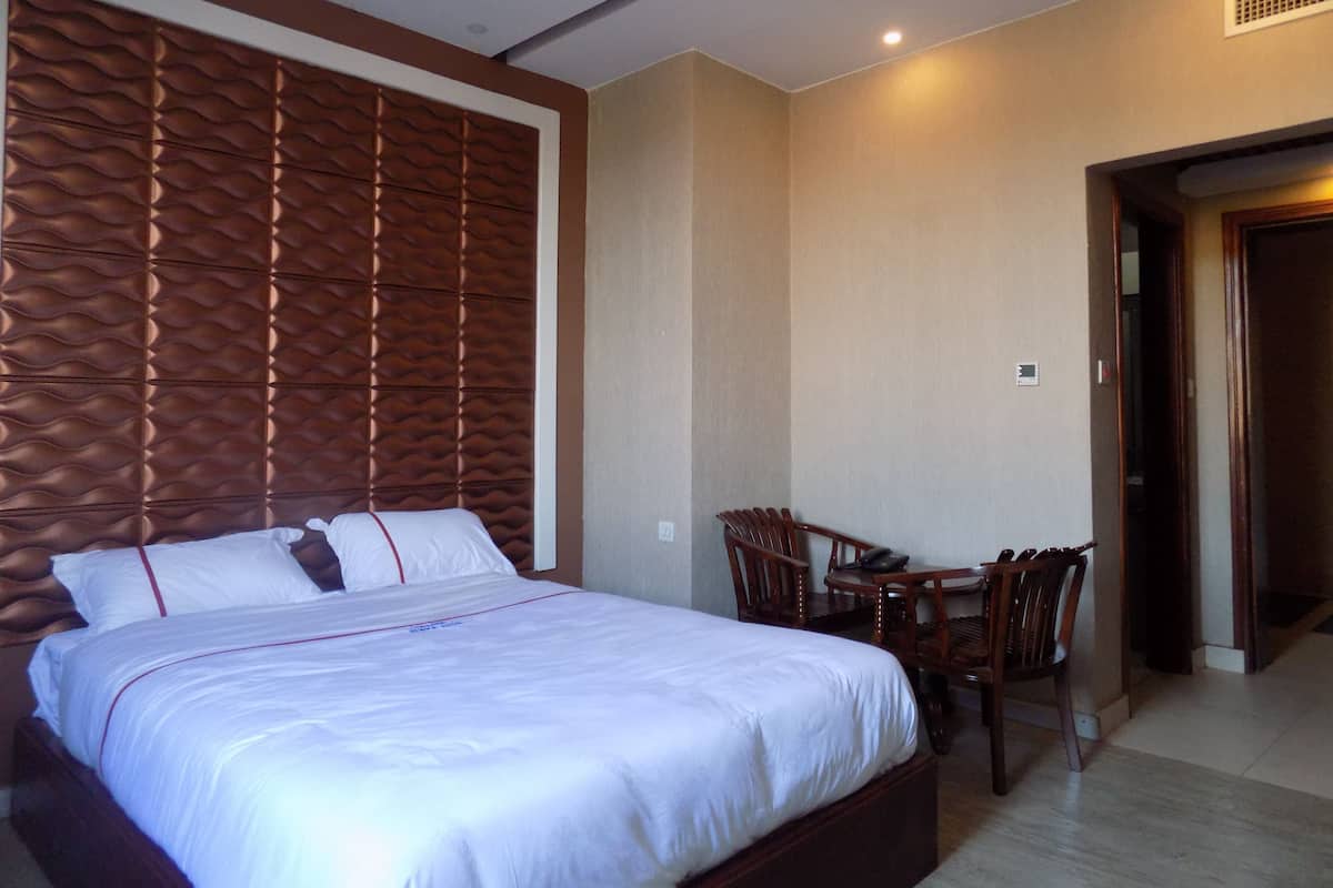 A hotel room with a large bed, a wooden headboard, a dining table with chairs, and a door leading to another room.