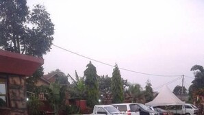 A paved parking area with several vehicles, including a white van and a blue car. There is a stone building with a red roof in the background. - Makutano Hotel (Kampala)