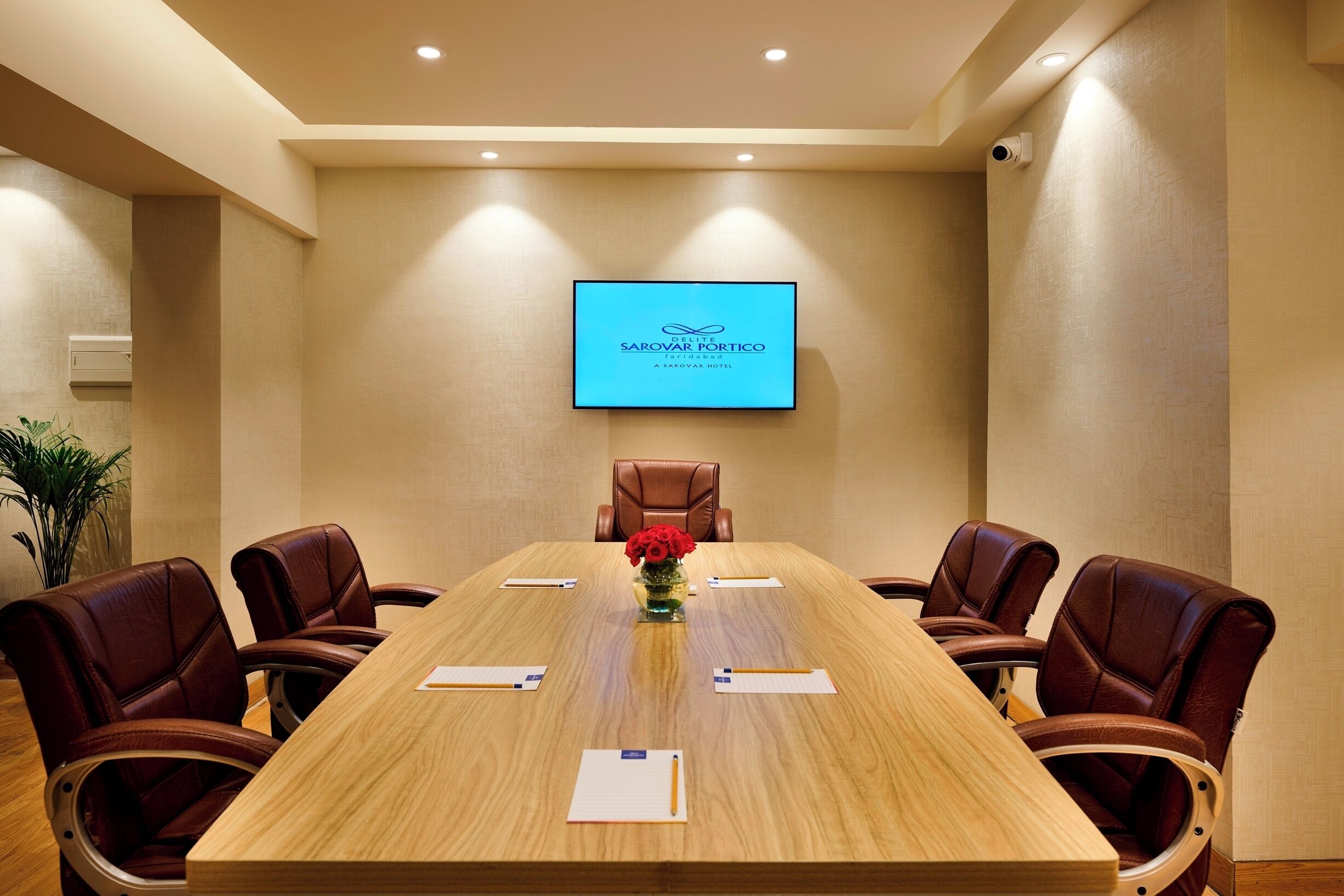 Ein Konferenzraum mit einem langen Tisch, sechs Ledersesseln, einer Vase mit roten Blumen und einem Bildschirm, der den Namen des Hotels zeigt.