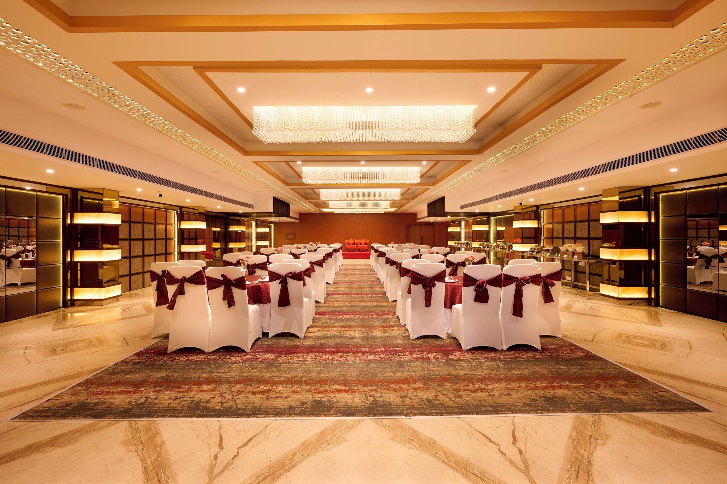 A spacious hotel lobby with a central aisle leading to a stage, surrounded by rows of white chairs with red sashes.