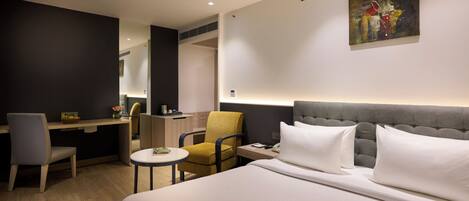 A hotel room with a bed, a desk, a chair, a small table, and a painting on the wall.