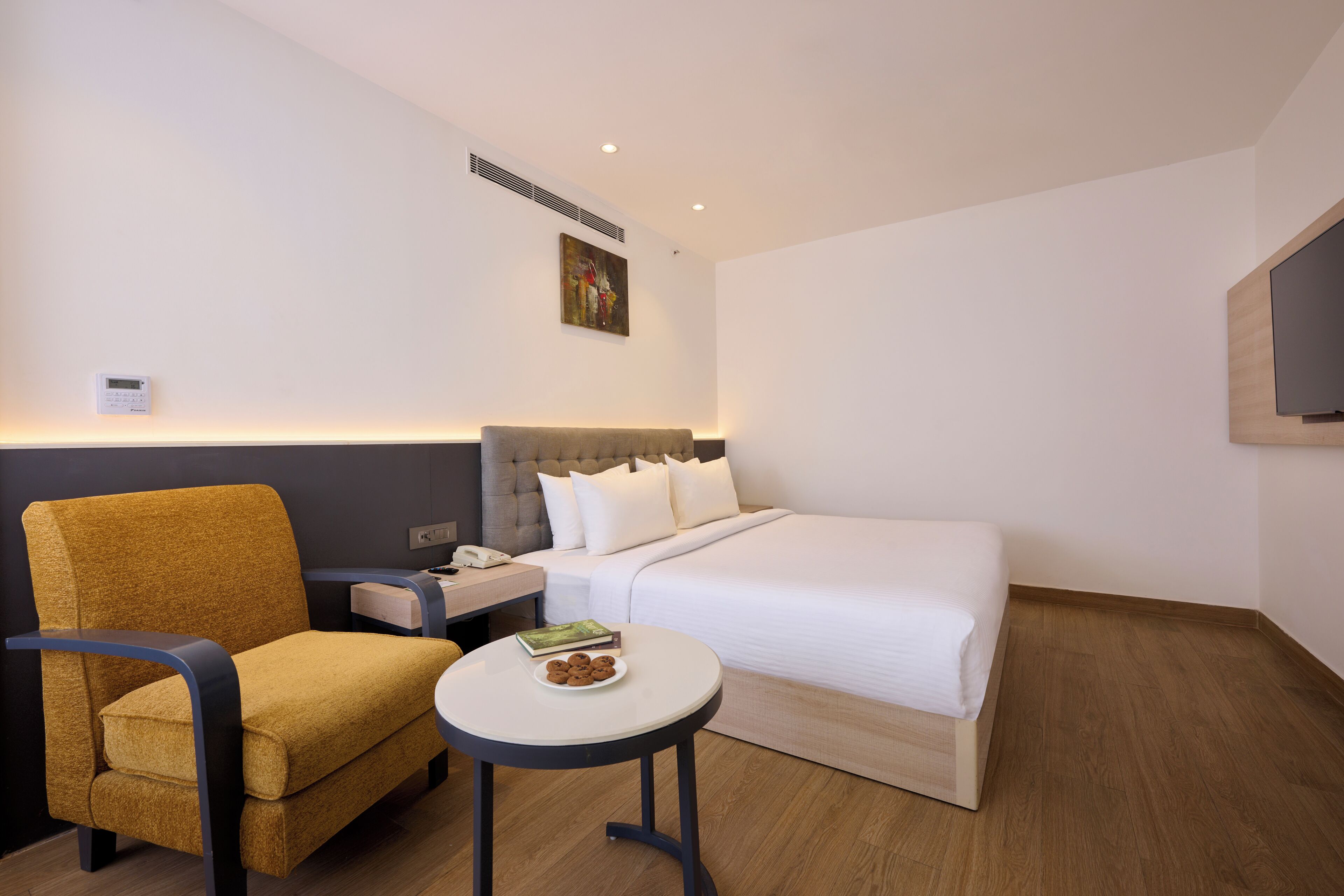 A hotel room with a bed, a chair, a small table with cookies, a nightstand, a phone, and a painting on the wall.