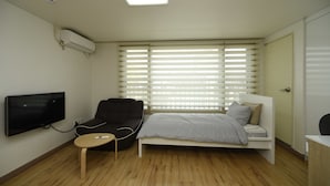 Ein modernes Schlafzimmer mit Bett, Fernseher, Sessel und einem Fenster mit Jalousien.