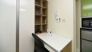 Ein kleiner, sauberer Raum mit weißem Schreibtisch, einem Stuhl, einem Bücherregal, einem Telefon und einem Fenster mit grünem Rahmen.