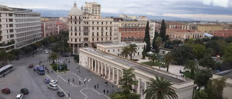 A cityscape with a prominent historical building featuring classical architecture, a busy intersection with vehicles and pedestrians, and a backdrop of other urban buildings.