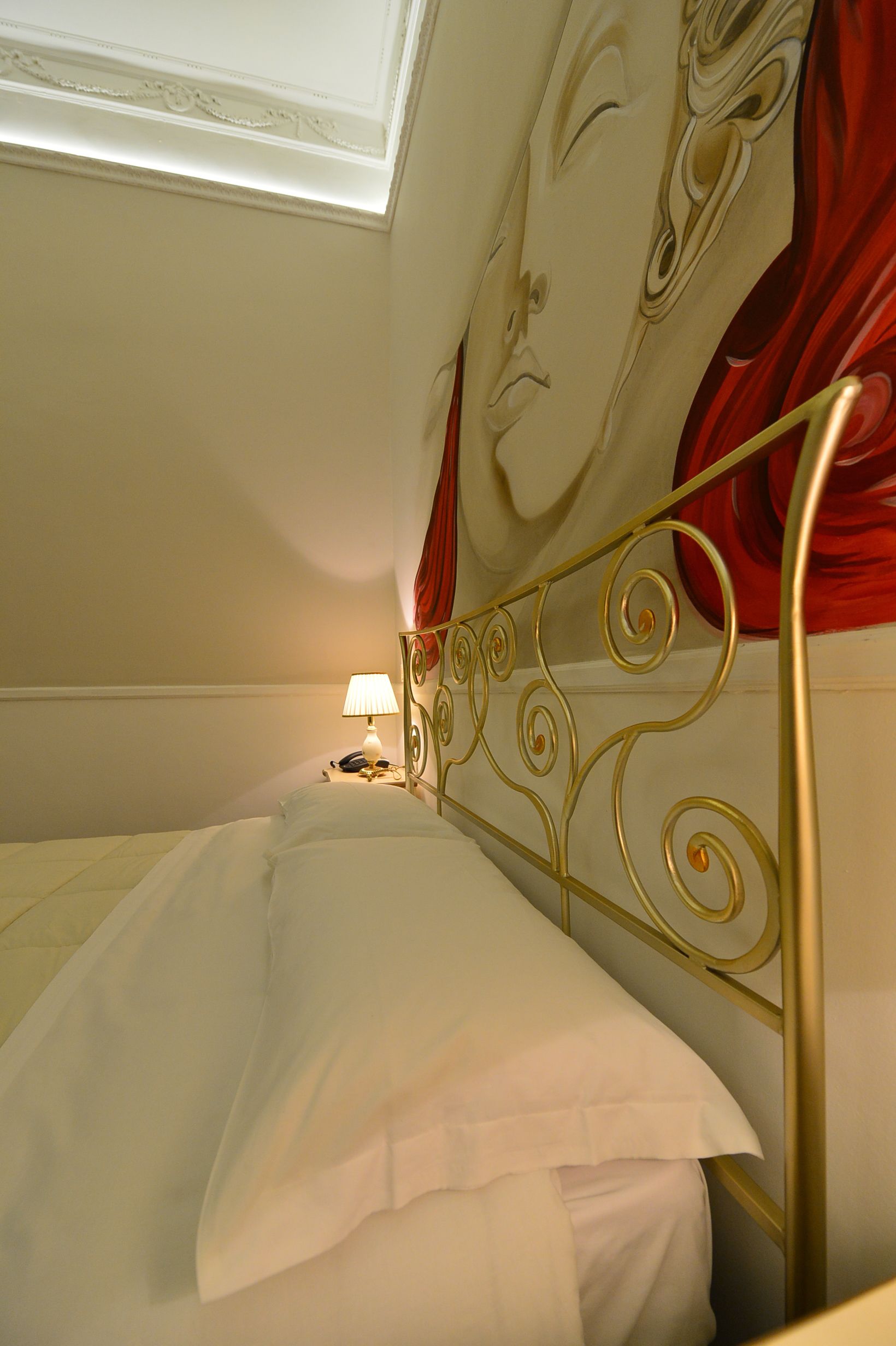 Un letto con lenzuola bianche, una testiera in metallo, una lampada da comodino e una parte di tenda rossa.