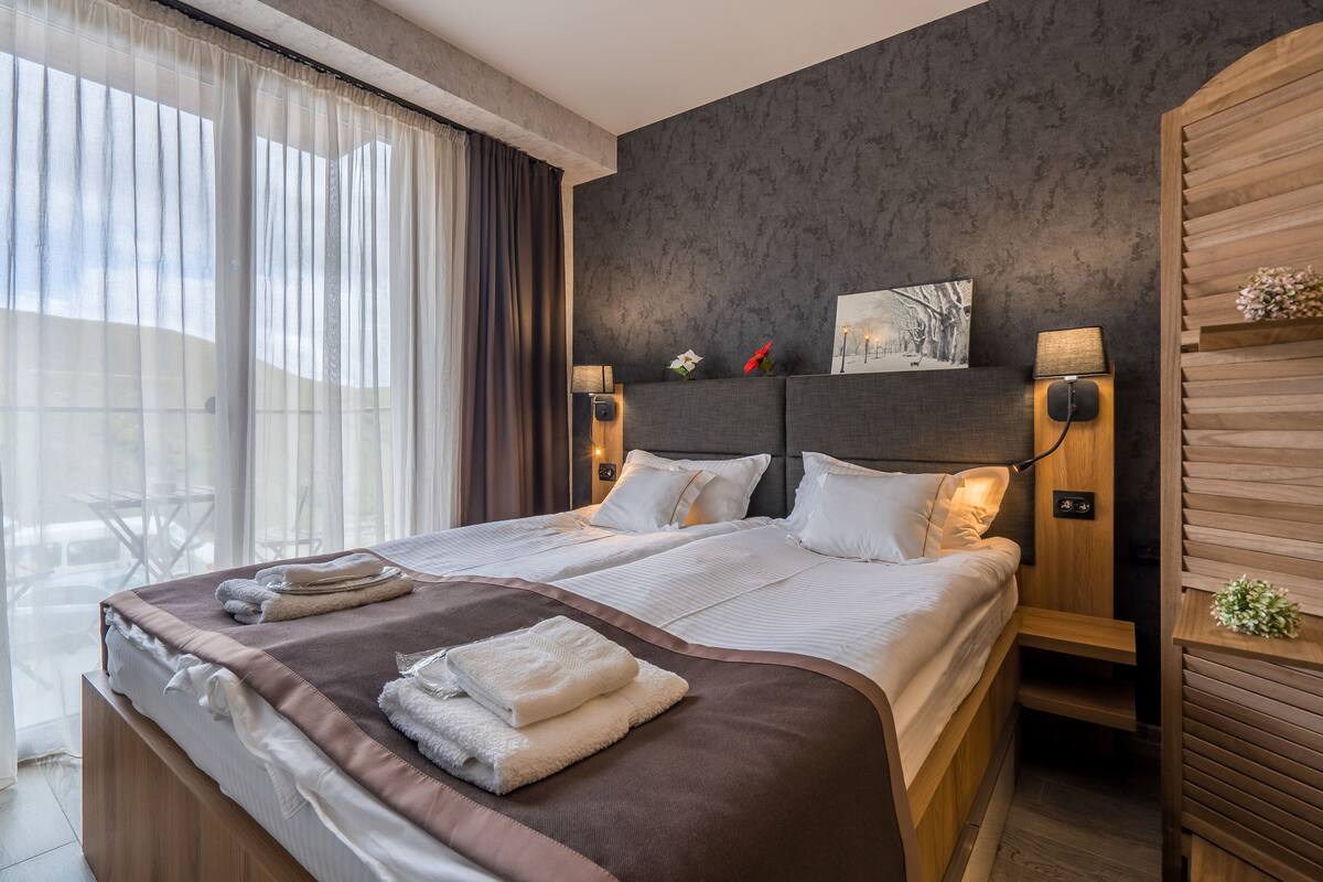 Una habitación de hotel moderna con una cama grande, dos mesitas de noche y vistas al exterior.
