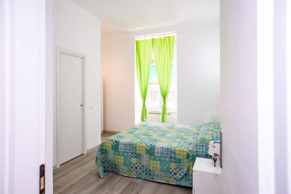 A small, clean room with a bed, a window with green curtains, and a white sink.