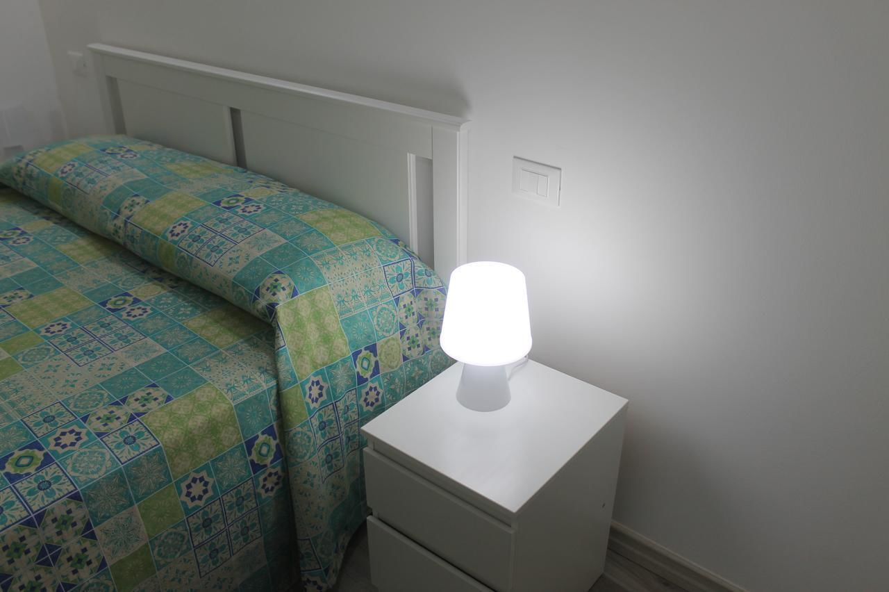 Ein Schlafzimmer mit einem Bett, das mit einer gemusterten Stegdecke bezogen ist, einem Nachttisch mit Lampe und einer weißen Wand mit Lichtschalter.
