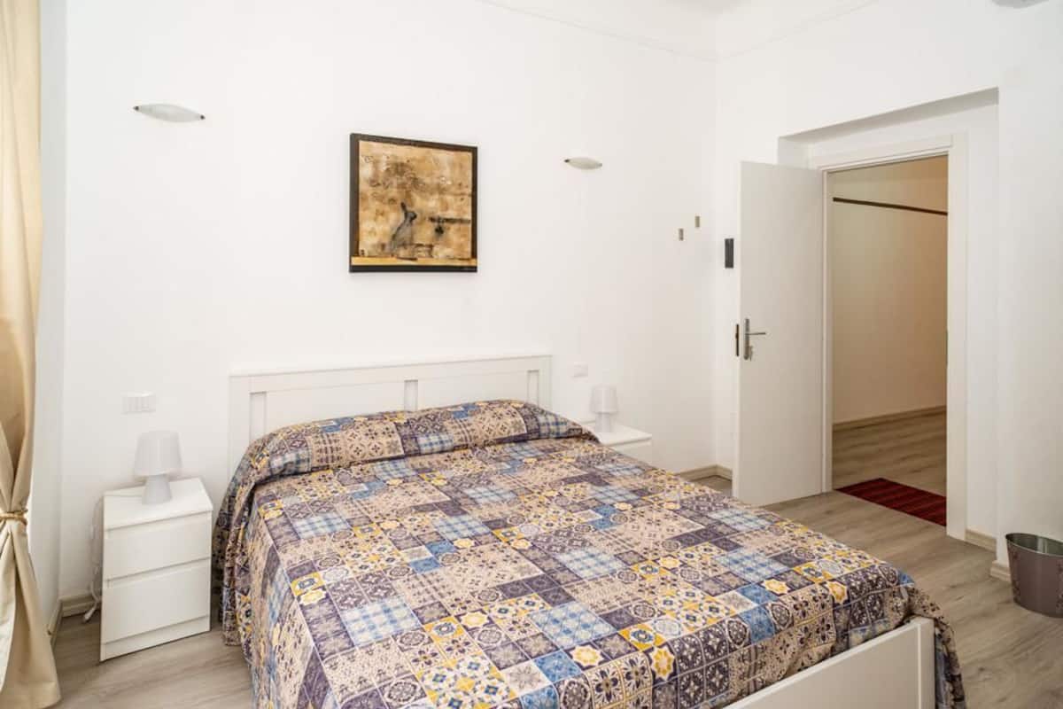 A bedroom with a patterned bedspread, a bedside table with a lamp, a wall-mounted artwork, and a door leading to another room.