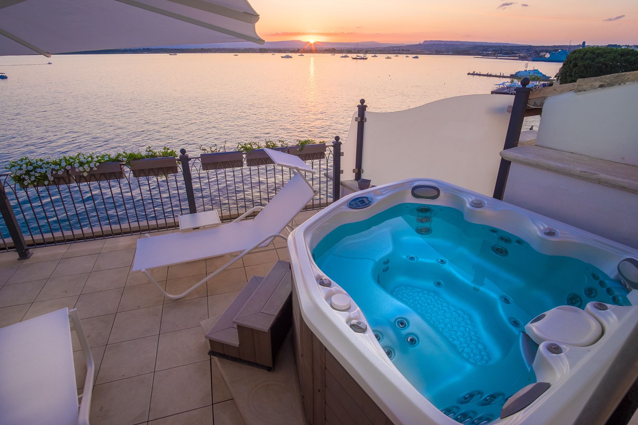 A balcony with a hot tub, lounge chairs, and a view of the sea at sunset.