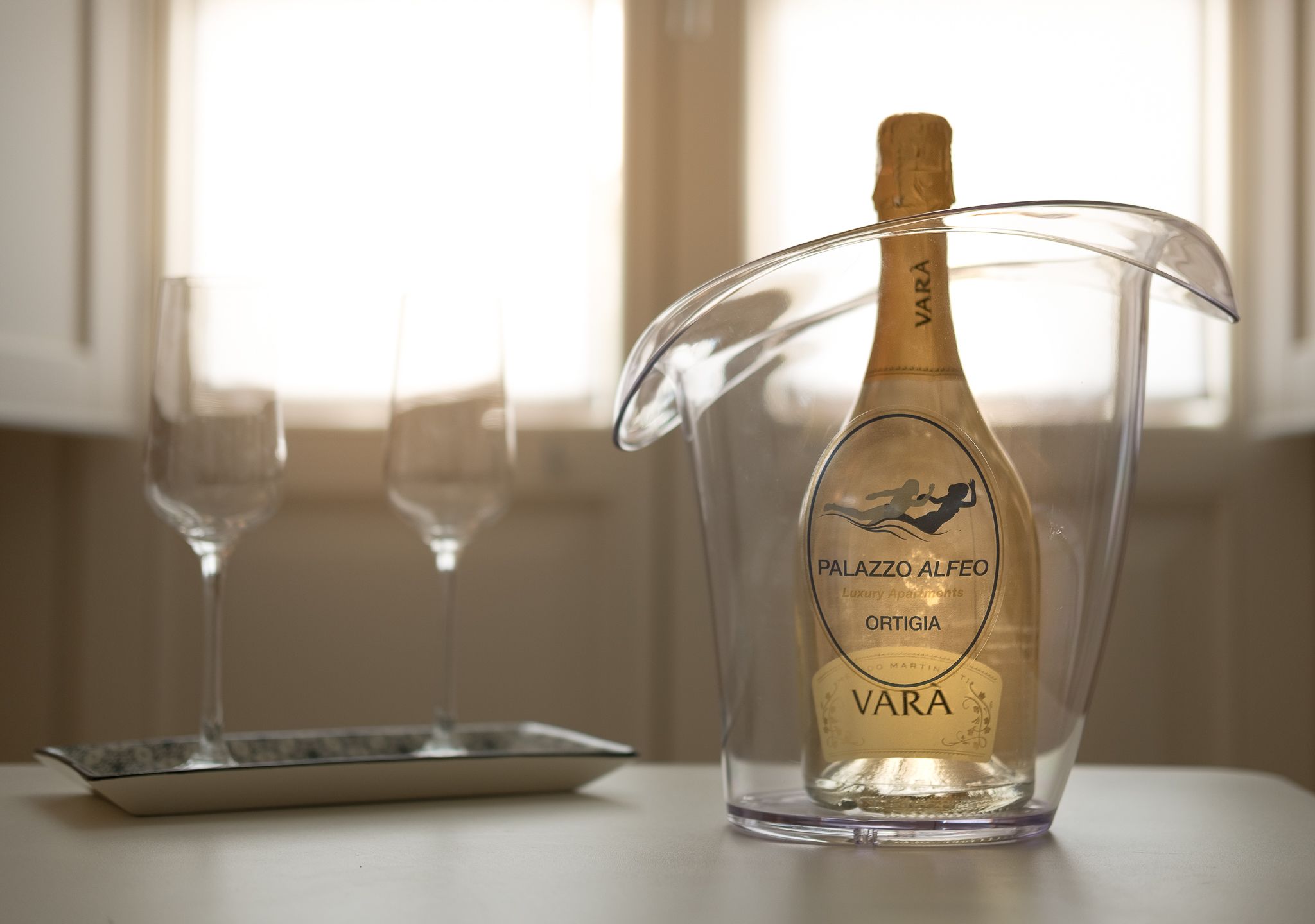Une bouteille de vin Palazzo Alfeo Ortigia VARA dans une carafe en verre transparent, posée sur une table avec deux verres à vin vides.