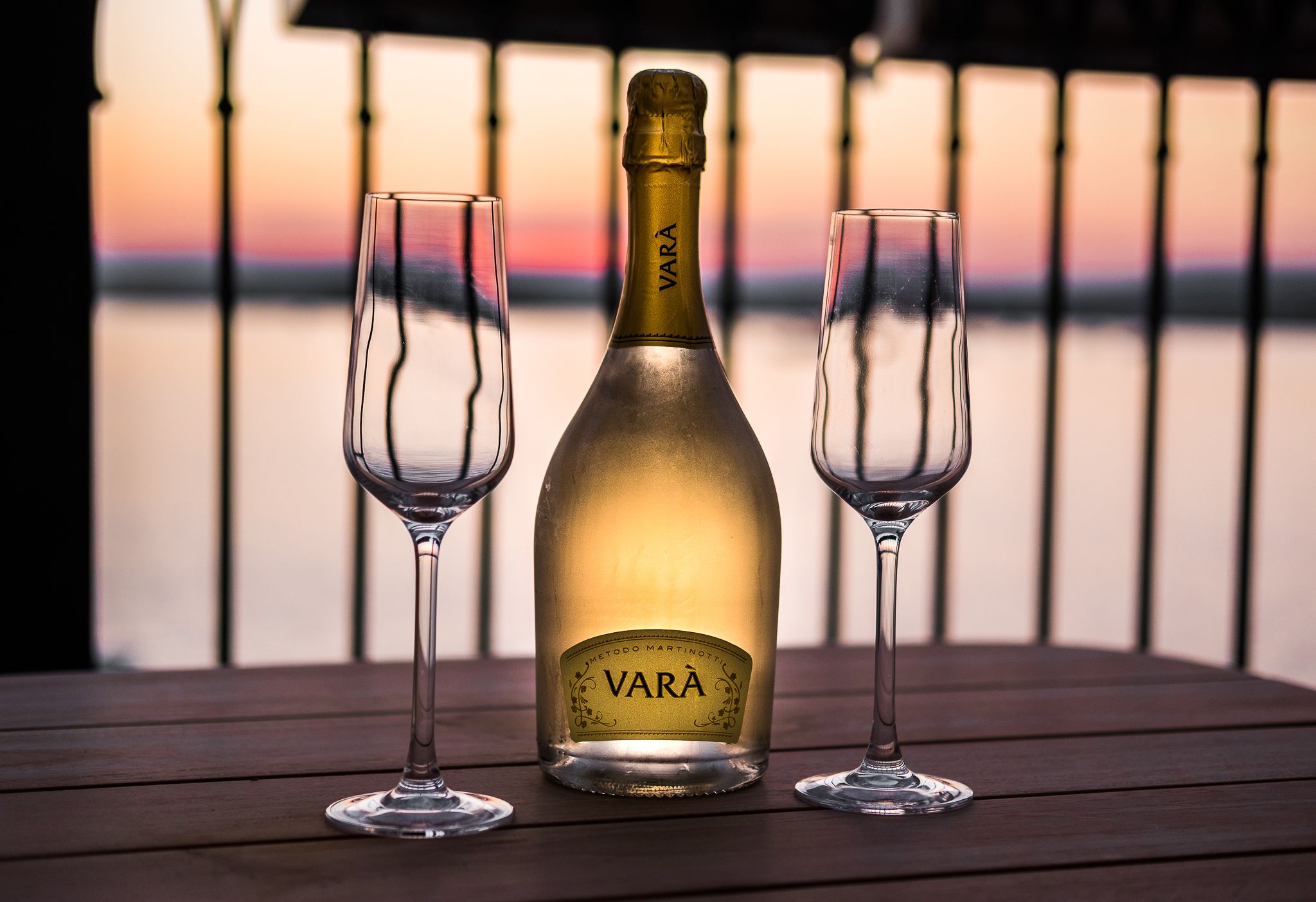 A bottle of VARA wine with two glasses on a wooden table against a sunset backdrop.
