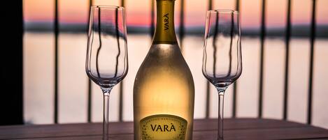 A bottle of VARA wine with two glasses on a wooden table against a sunset backdrop.