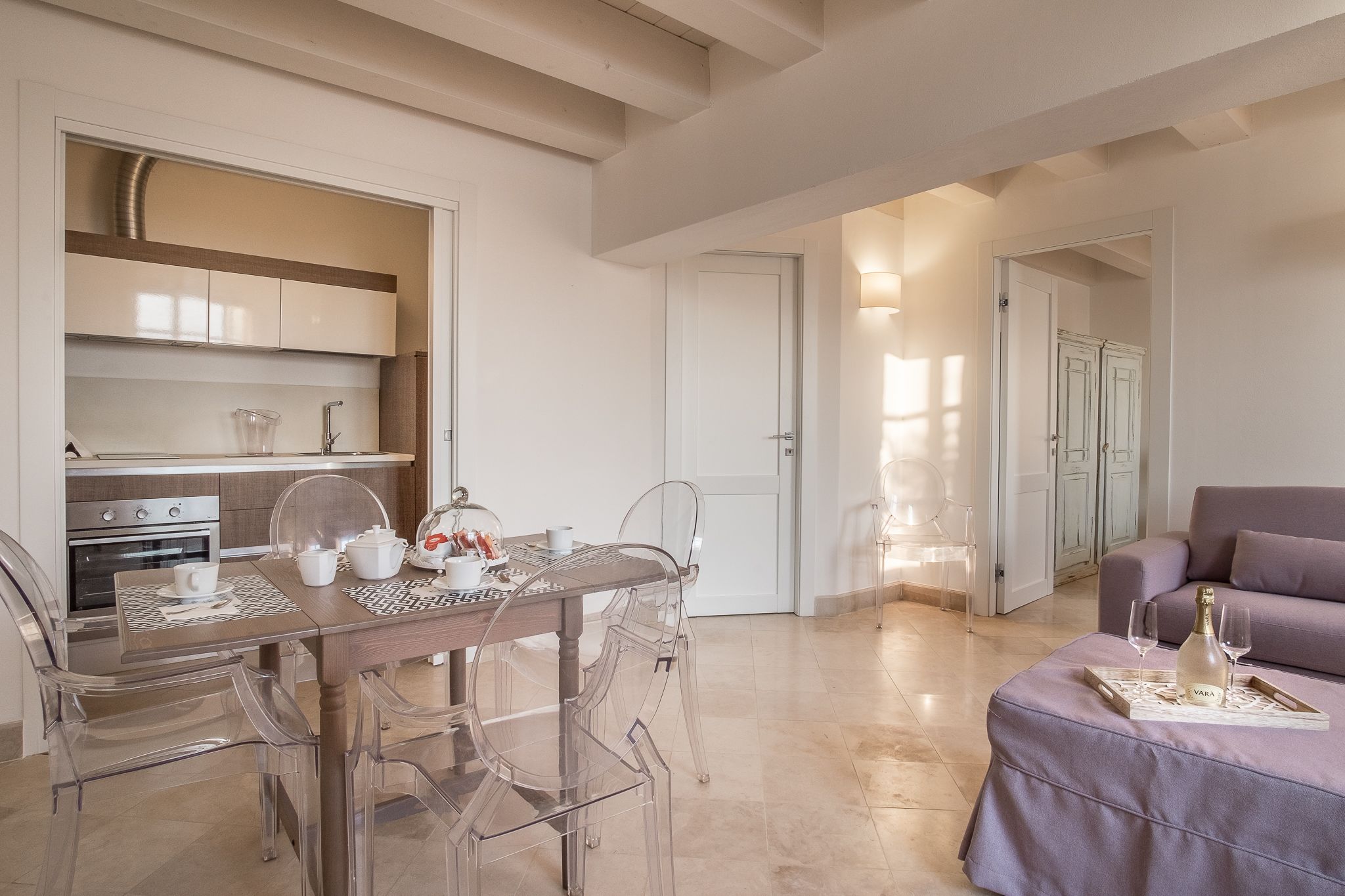 A modern kitchen with stainless steel appliances, a dining area with clear chairs, and a living area with a sofa and a tray table.