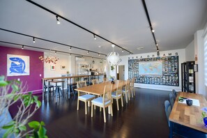 A modern dining area with wooden tables and chairs, a kitchen in the background, and a wall with a map and various items. - Ise Guest House sora - Hostel (Ise)