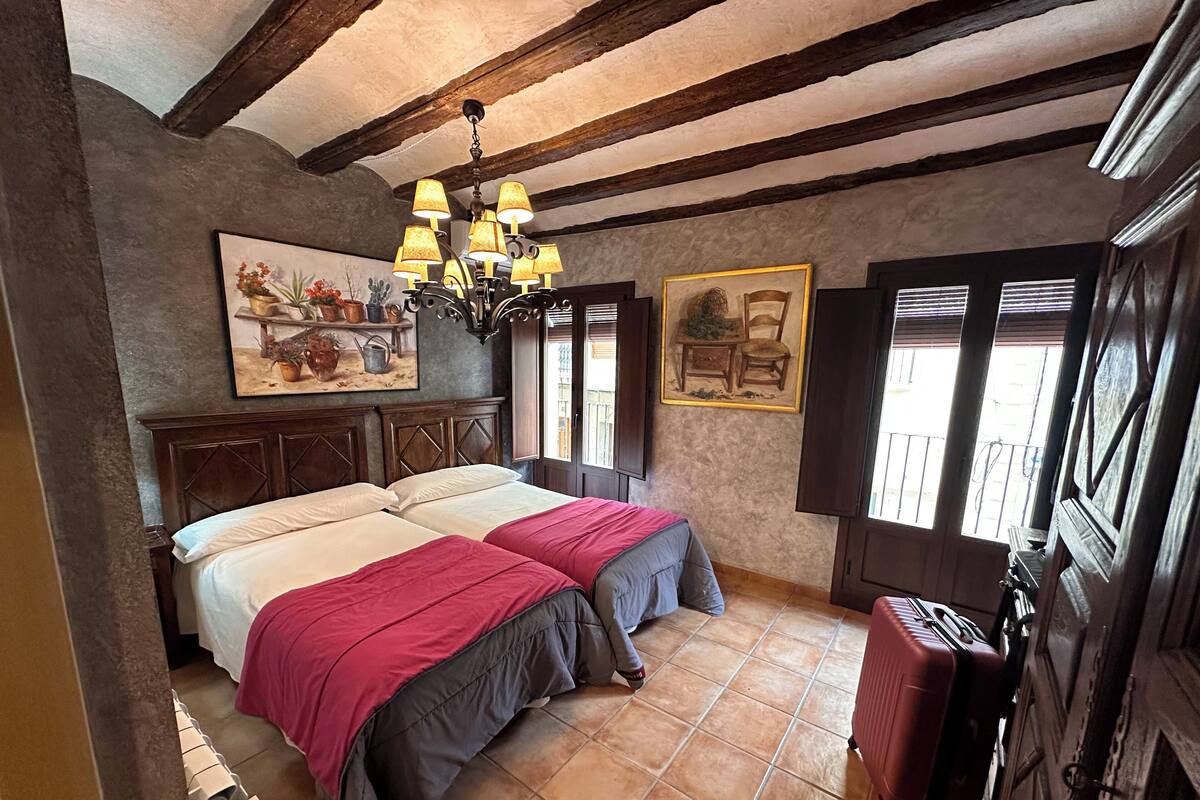 A bedroom with two beds, a chandelier, a painting, and a suitcase.