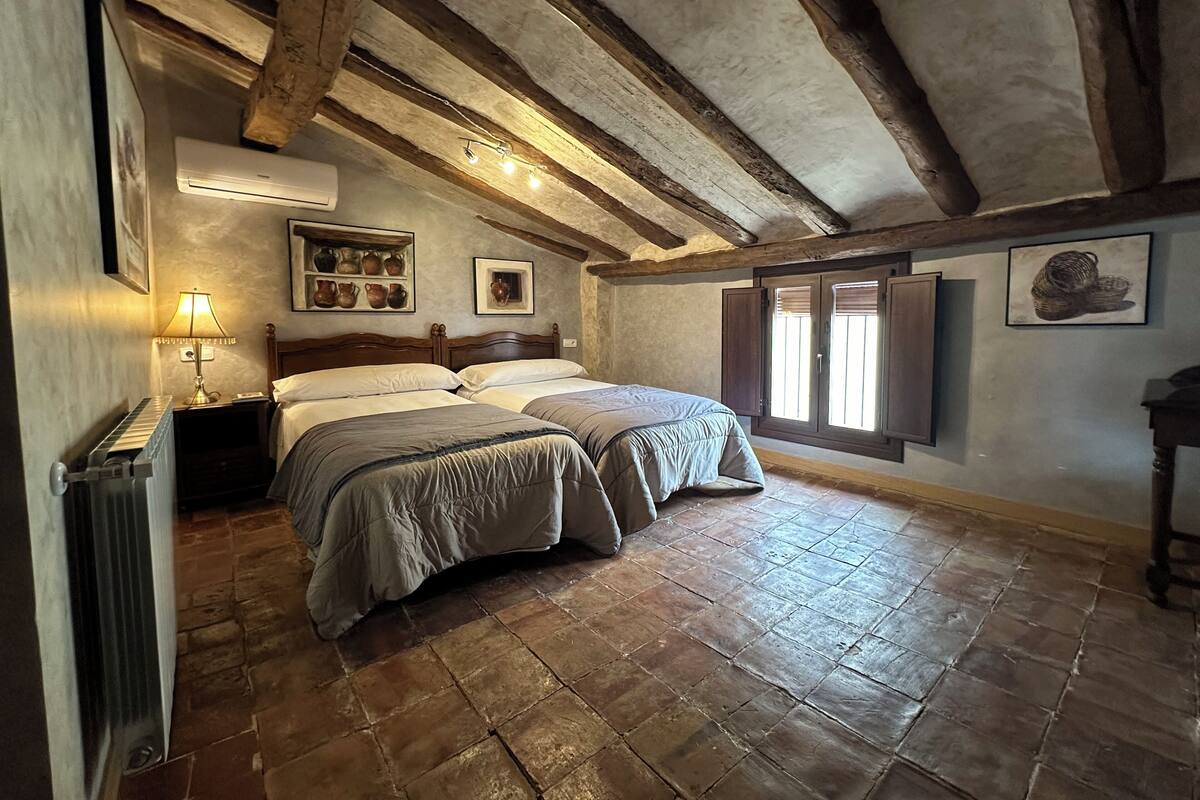 A rustic bedroom with two beds, a radiator, a lamp, and a shelf with pottery.