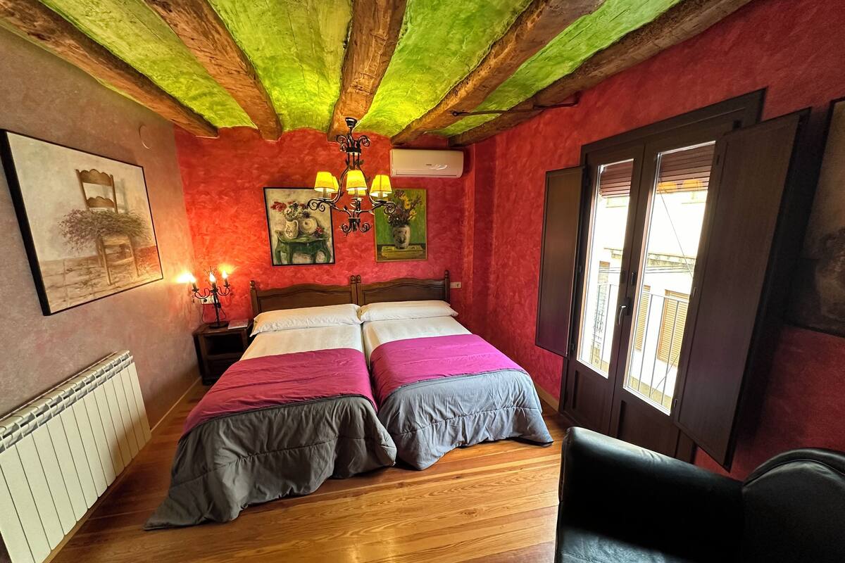 A room with a wooden floor, a bed with pink and grey bedding, a black leather chair, a chandelier, and artwork on the walls.