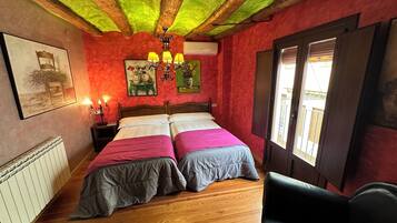 Una habitación con piso de madera, una cama con ropa de cama rosa y gris, un sillón de cuero negro, un candelabro y obras de arte en las paredes.