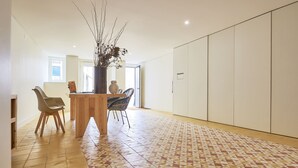 Una habitación amplia con suelo de baldosas, una mesa de comedor de madera con sillas, un jarrón con ramas y un armario.