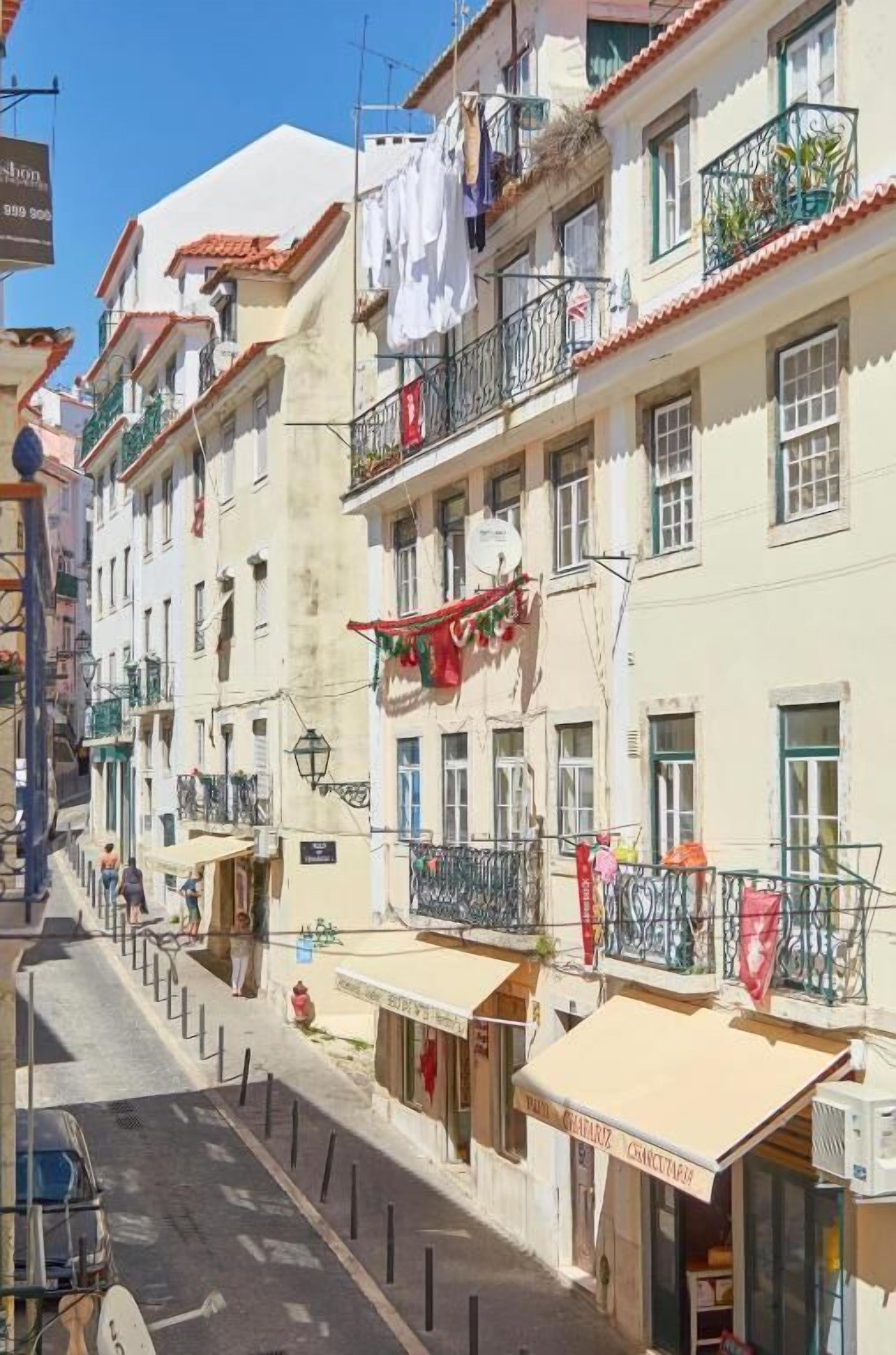 A narrow street lined with multi-story buildings, balconies with plants, and a few pedestrians.