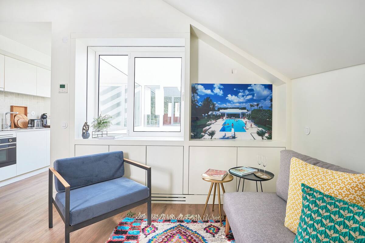 A modern living room with a sofa, a blue chair, a small table with a book and glasses, and a colorful rug.