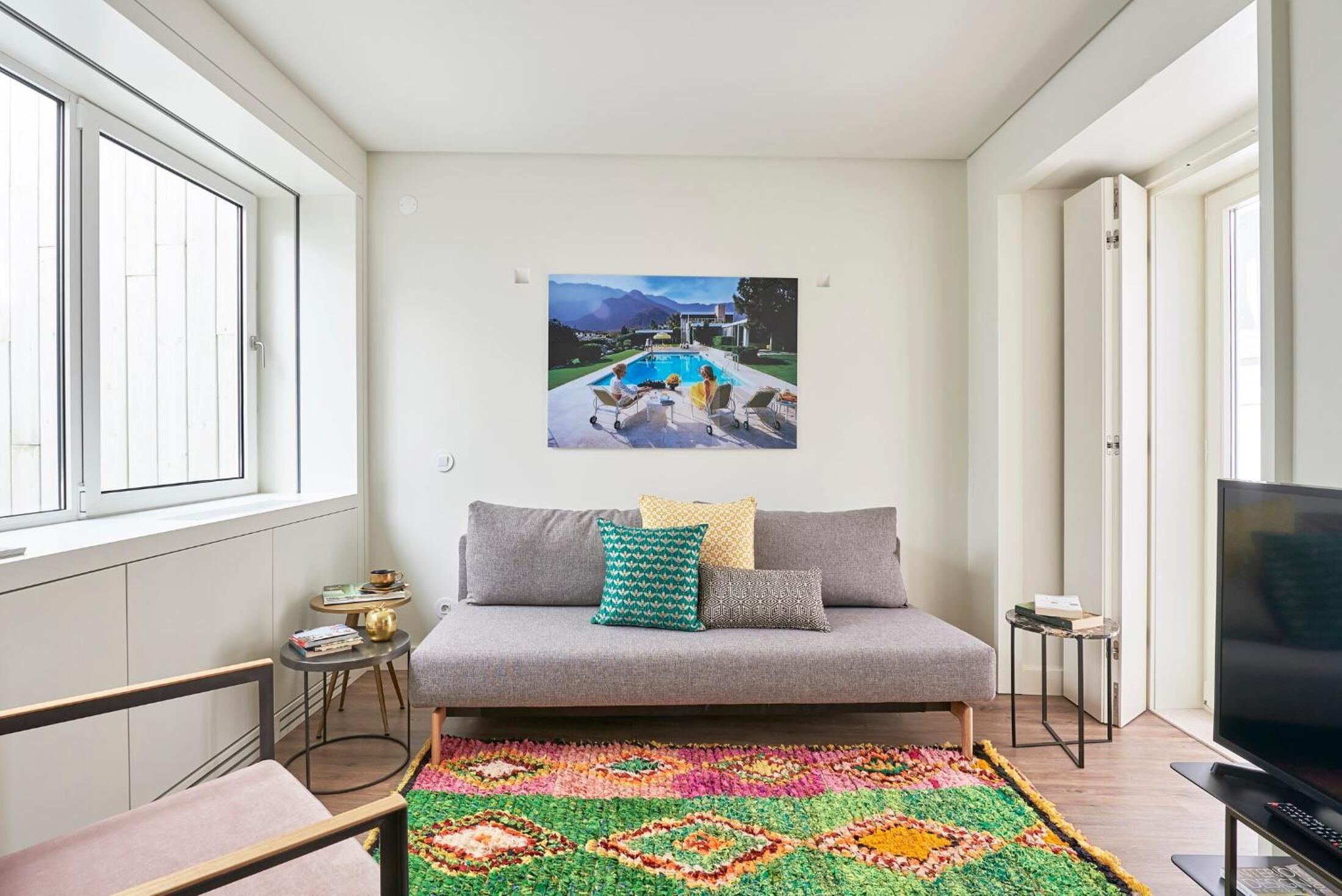 Ein modernes Wohnzimmer mit grauem Sofa, farbenfrohem Teppich und einem Gemälde, das eine Poolszene zeigt.