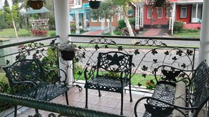 A covered outdoor seating area with wrought iron chairs and a bench, overlooking a garden and buildings in the background. - Kota Bunga Q (Cipanas)