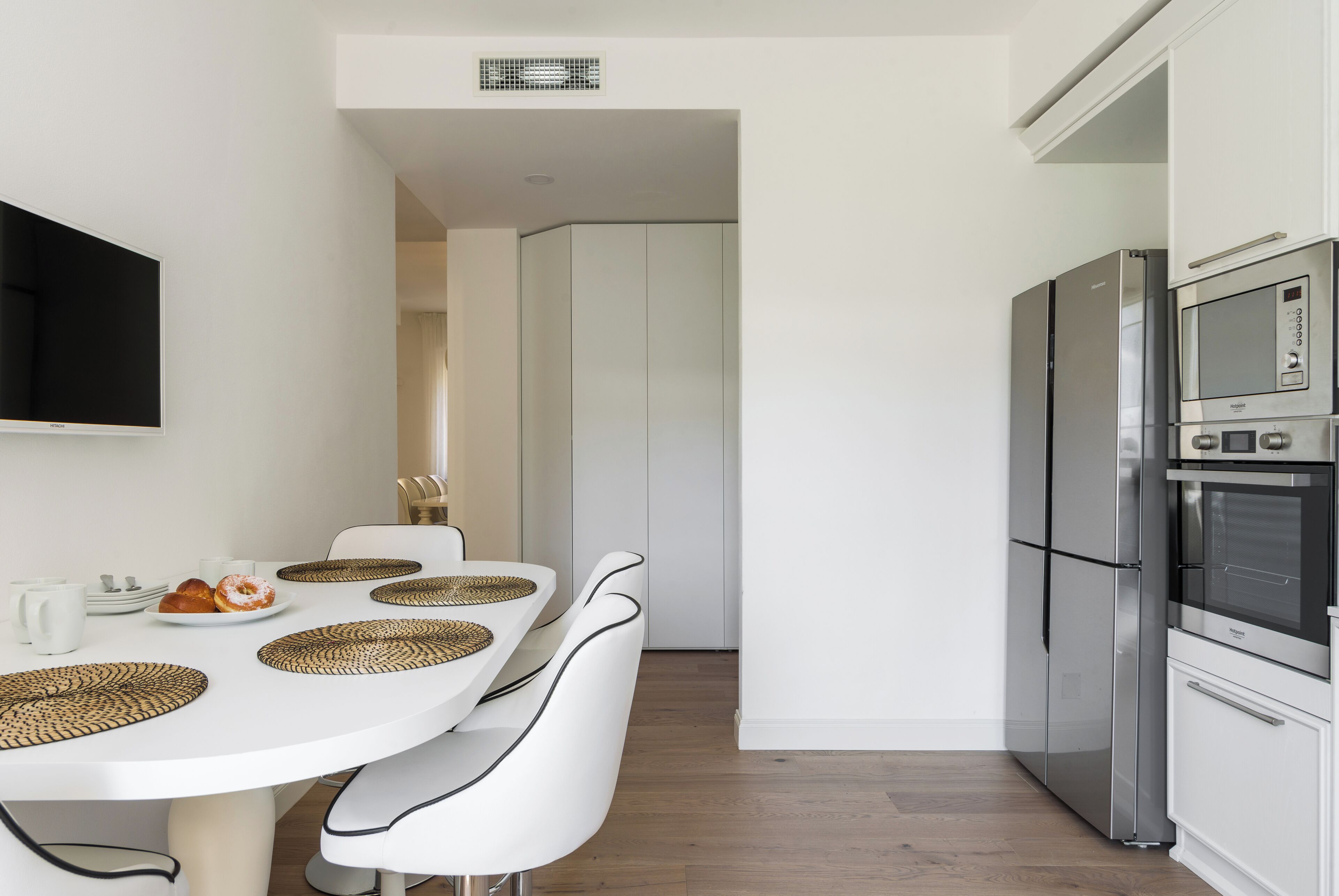 Eine moderne Küche mit Edelstahlgeräten, einem weißen Esstisch mit Platzdeckchen und einem an der Wand befestigten Flachbildfernseher.