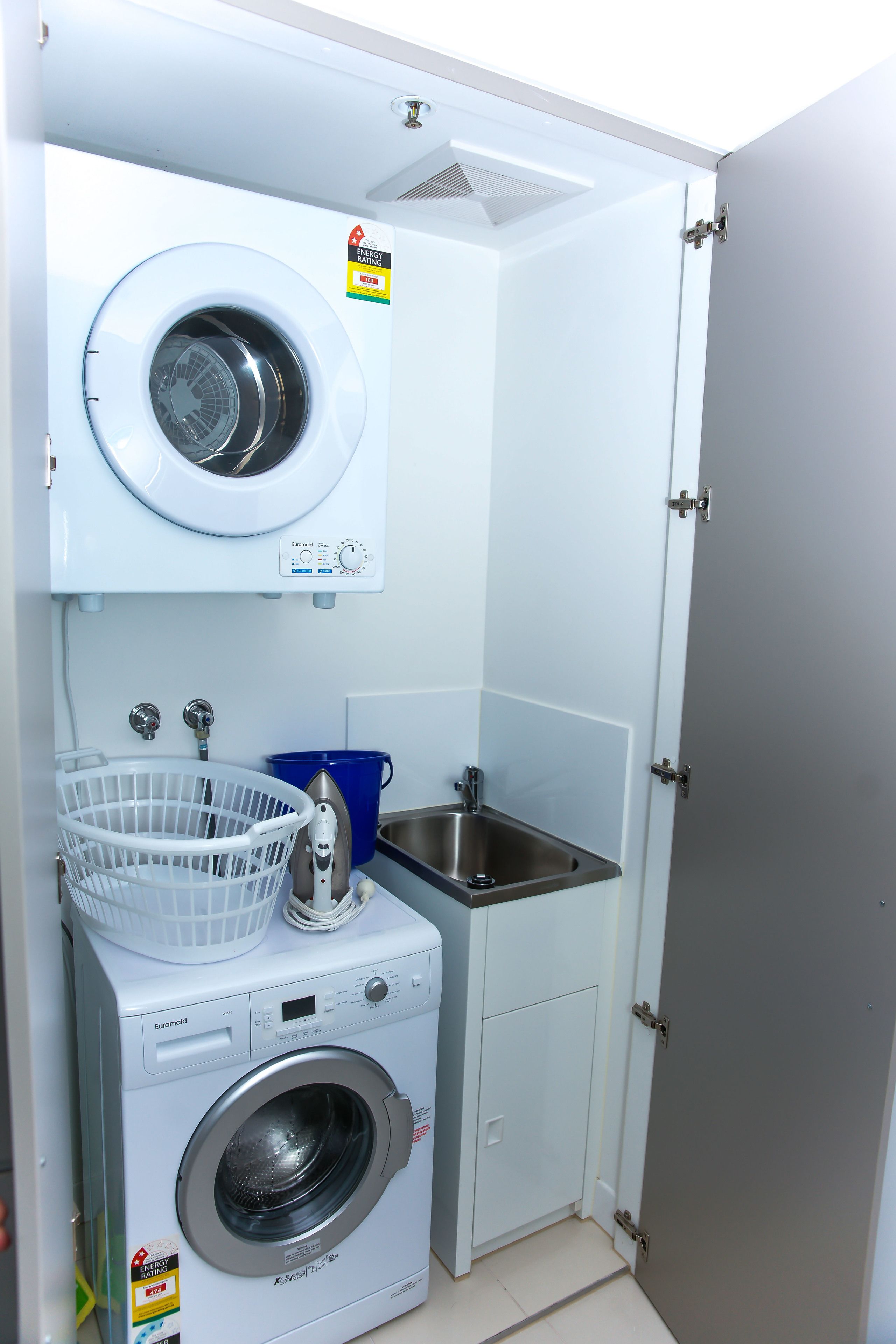 A compact laundry room with a front-loading washing machine, a small sink, and a blue bucket.