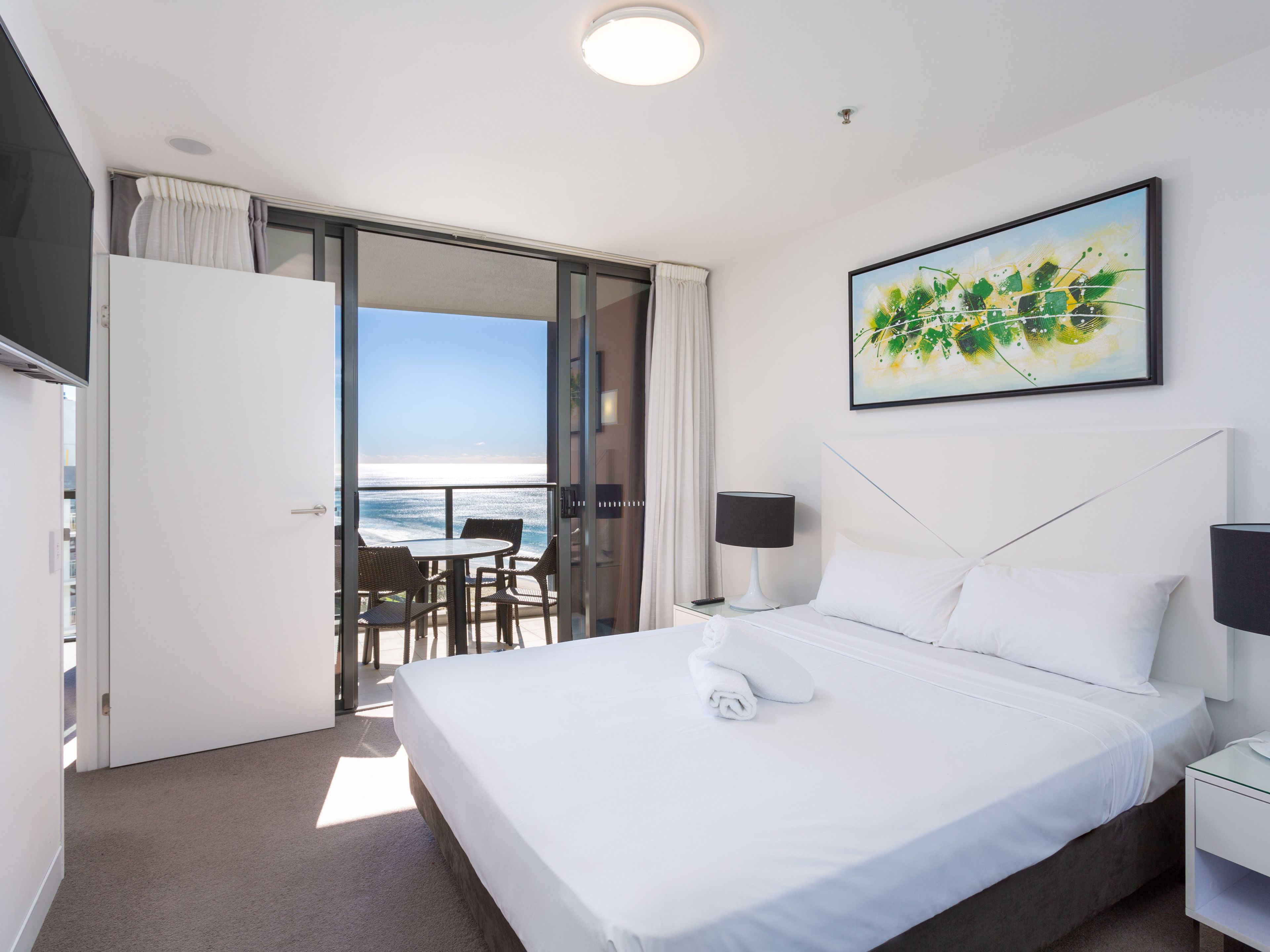 A hotel room with a bed, a painting, a nightstand, a lamp, and a view of the ocean.