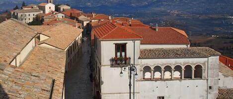 Una calle estrecha con edificios tradicionales, una torre de campanas y vistas a colinas distantes.